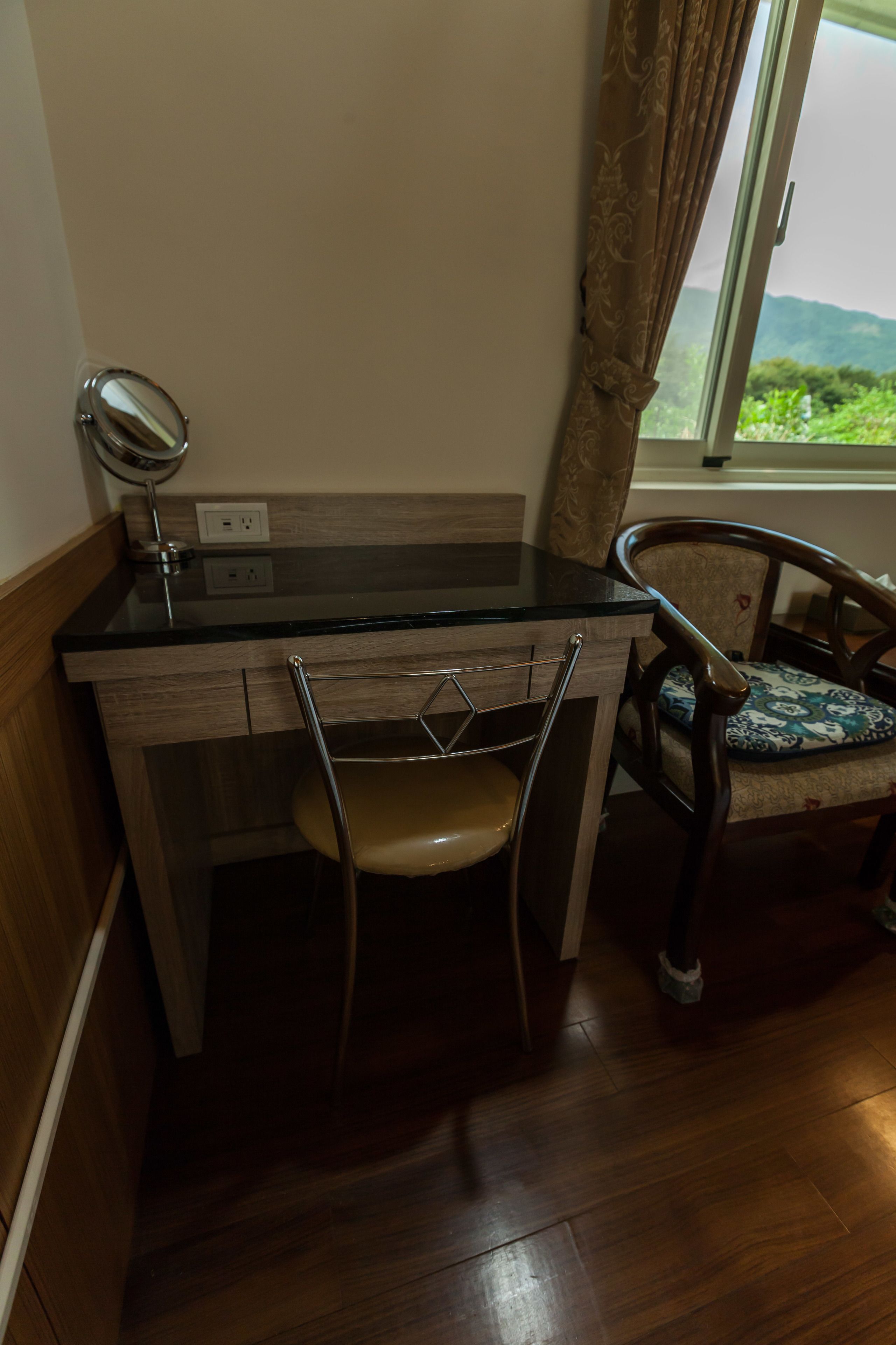 A compact room with a wooden desk, a chair, a mirror, and a window with a view of greenery.