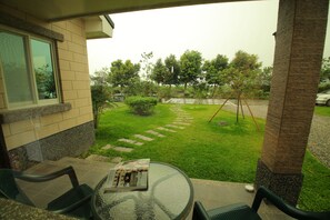 A patio area with a glass table, two chairs, and a magazine. There is a garden with a lawn, stepping stones, and trees.