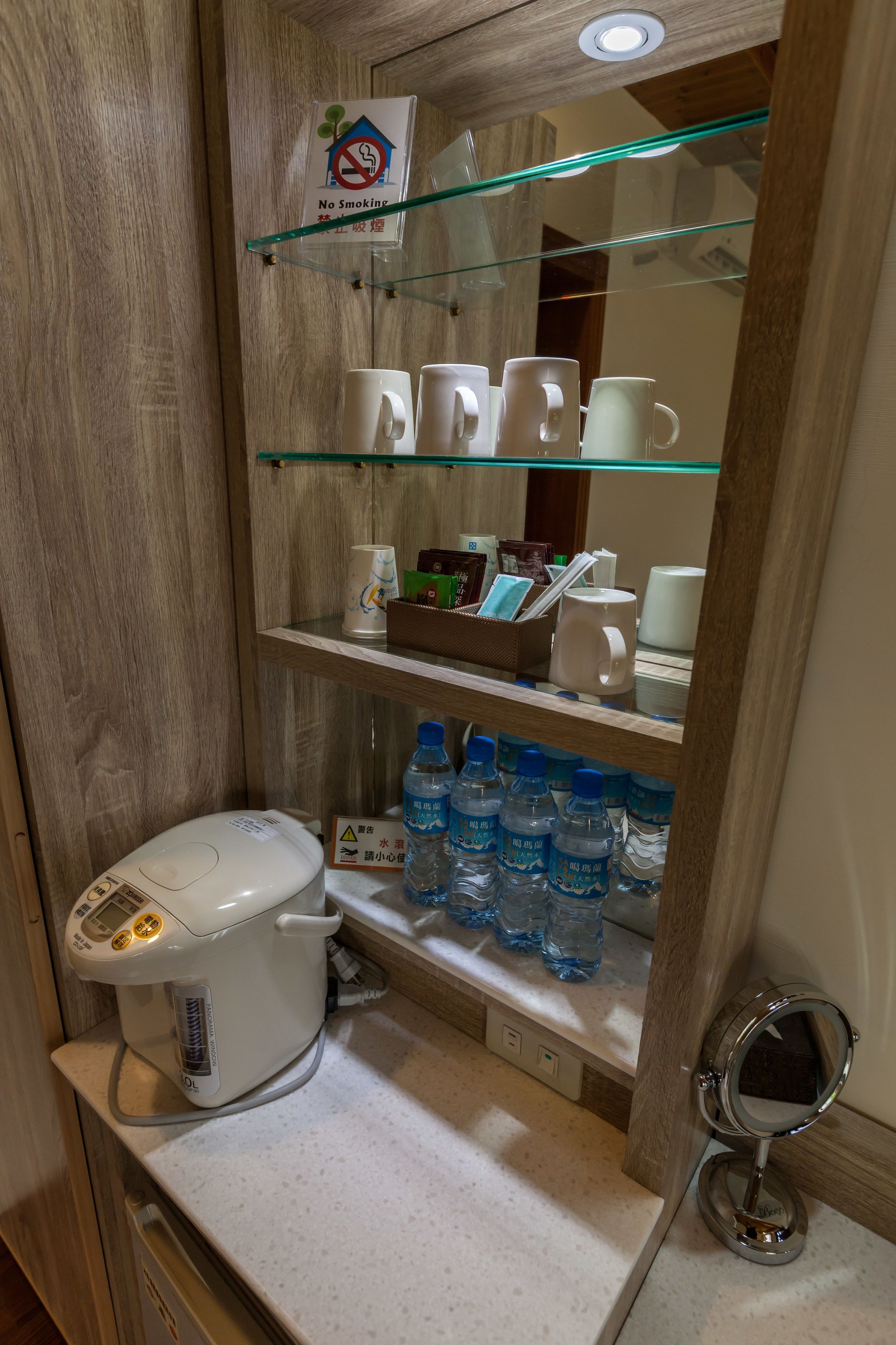 Un baño pequeño con inodoro blanco, repisa de vidrio con artículos de aseo, dispensador de agua y letrero de no fumar.