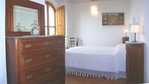 A bedroom with a wooden dresser, a bed with a white bedspread, a nightstand with a lamp, and a painting on the wall.