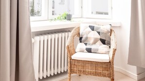 A bright room with a wicker chair, a white radiator, and a view of buildings through a large window. -  2ndhomes Beautiful & Modern 1BR Home with Central Location (Helsinki)