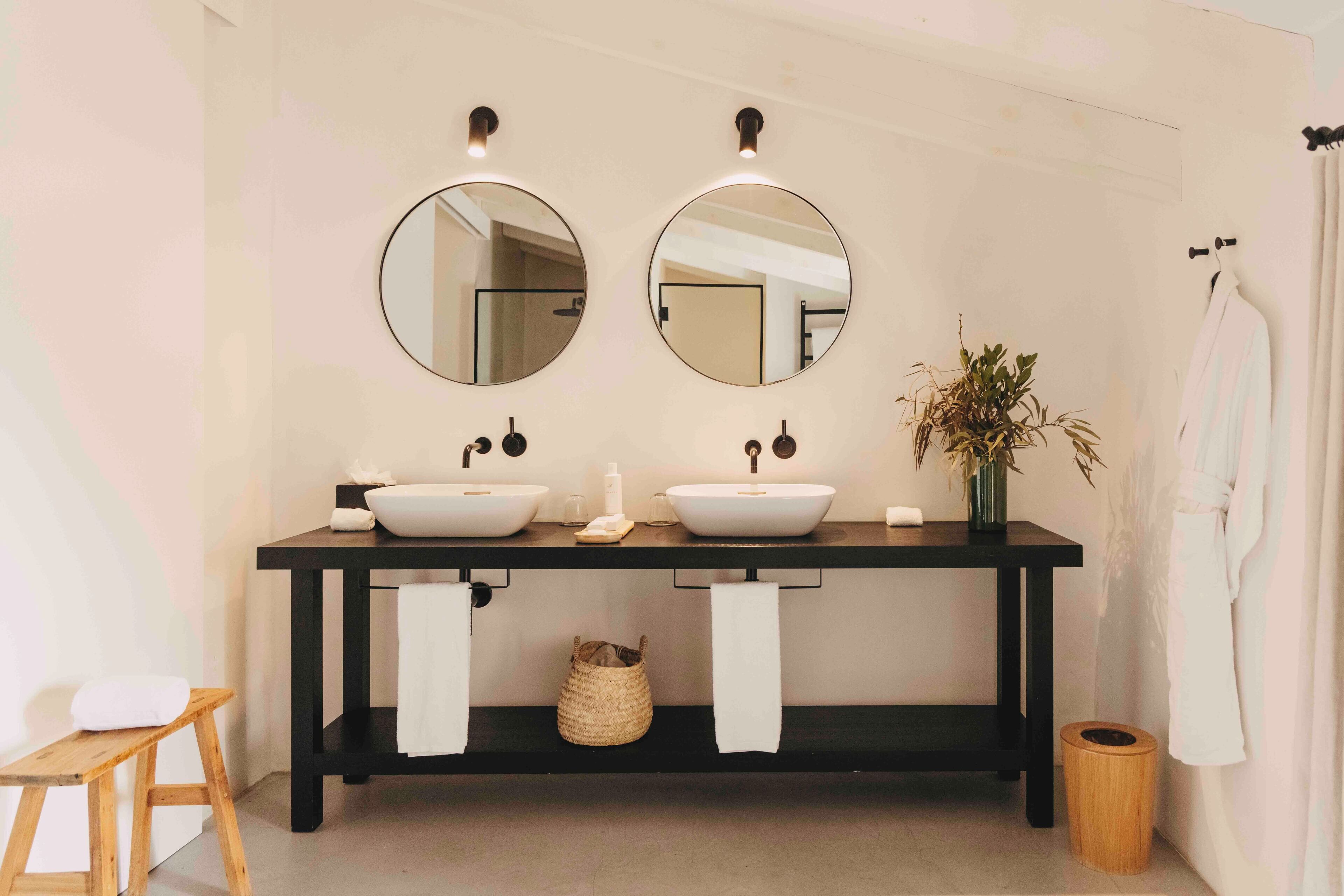 Un baño con dos espejos redondos, un mueble de tocador de madera oscura, dos lavamanos blancos y una planta pequeña.