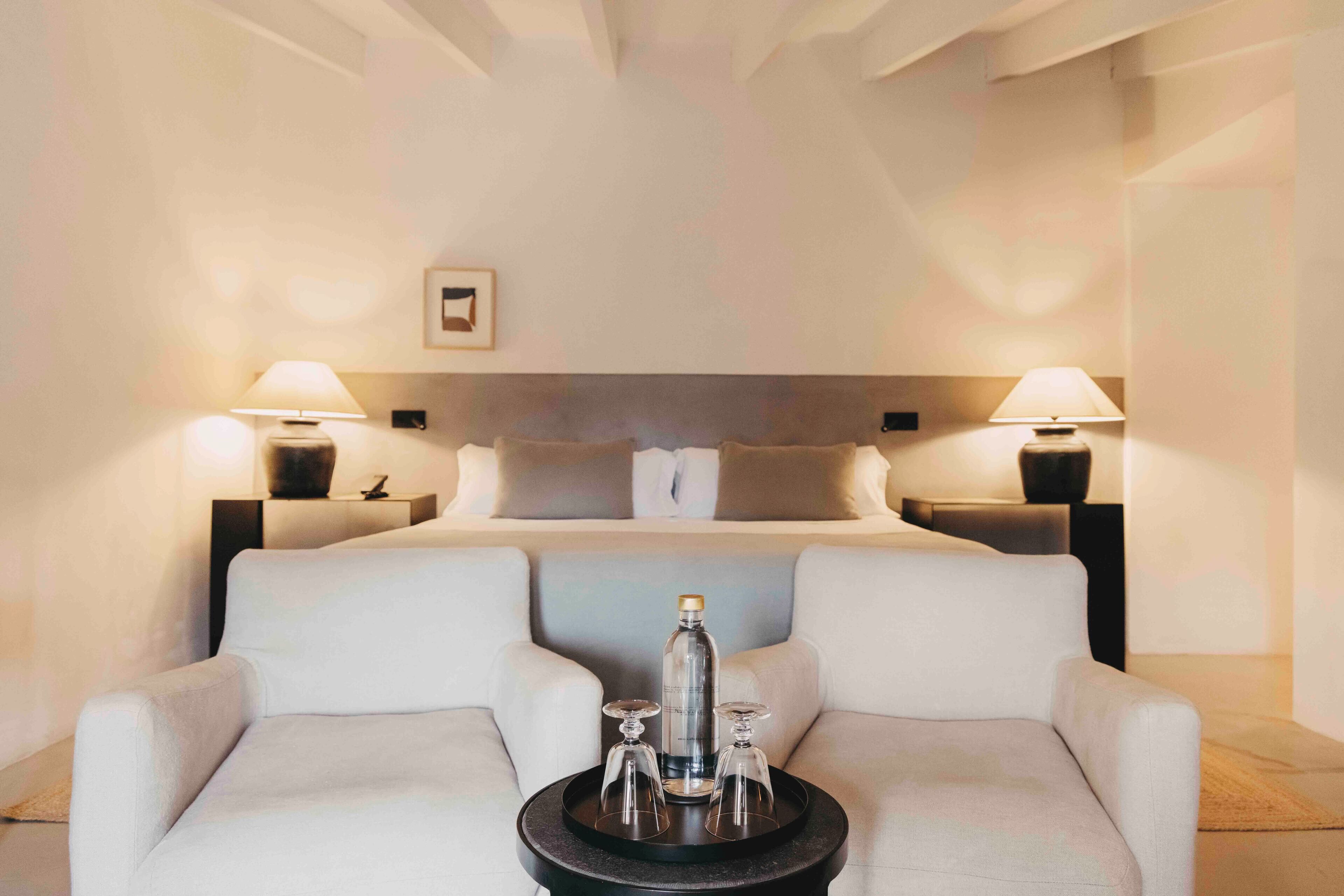 Una habitación de hotel moderna con una cama grande, dos mesitas de noche con lámparas, una pequeña mesa con una botella y vasos, y un sofá.