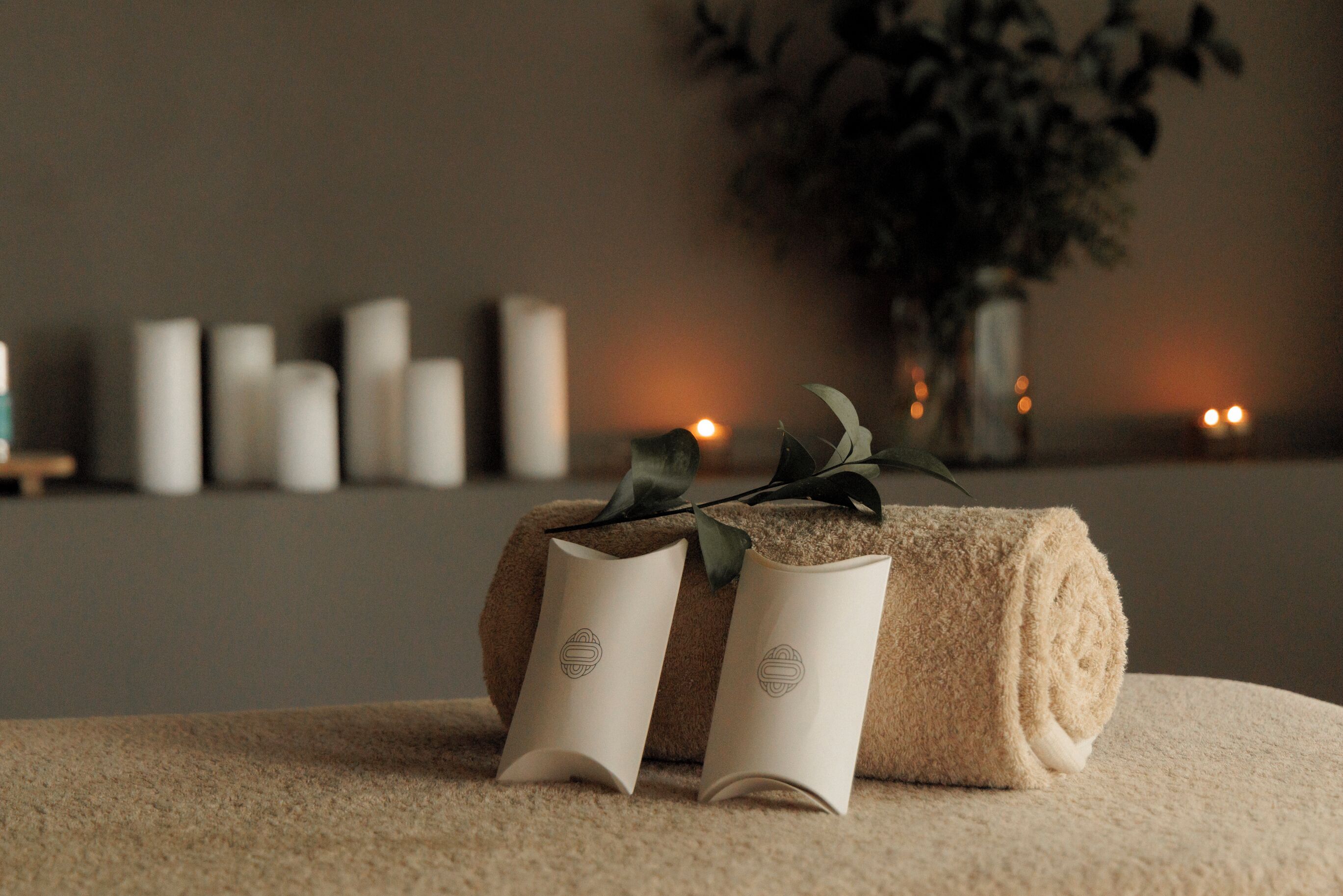 A neatly arranged room with a rolled beige towel and two folded towels on a textured surface, a vase with greenery, and a row of candles in the background.