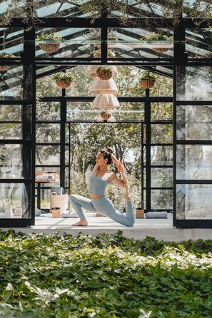 A person performing a yoga pose in a glass-enclosed pavilion with hanging planters and a view of trees. - Finca Serena Mallorca, Small Luxury Hotels (Montuiri)