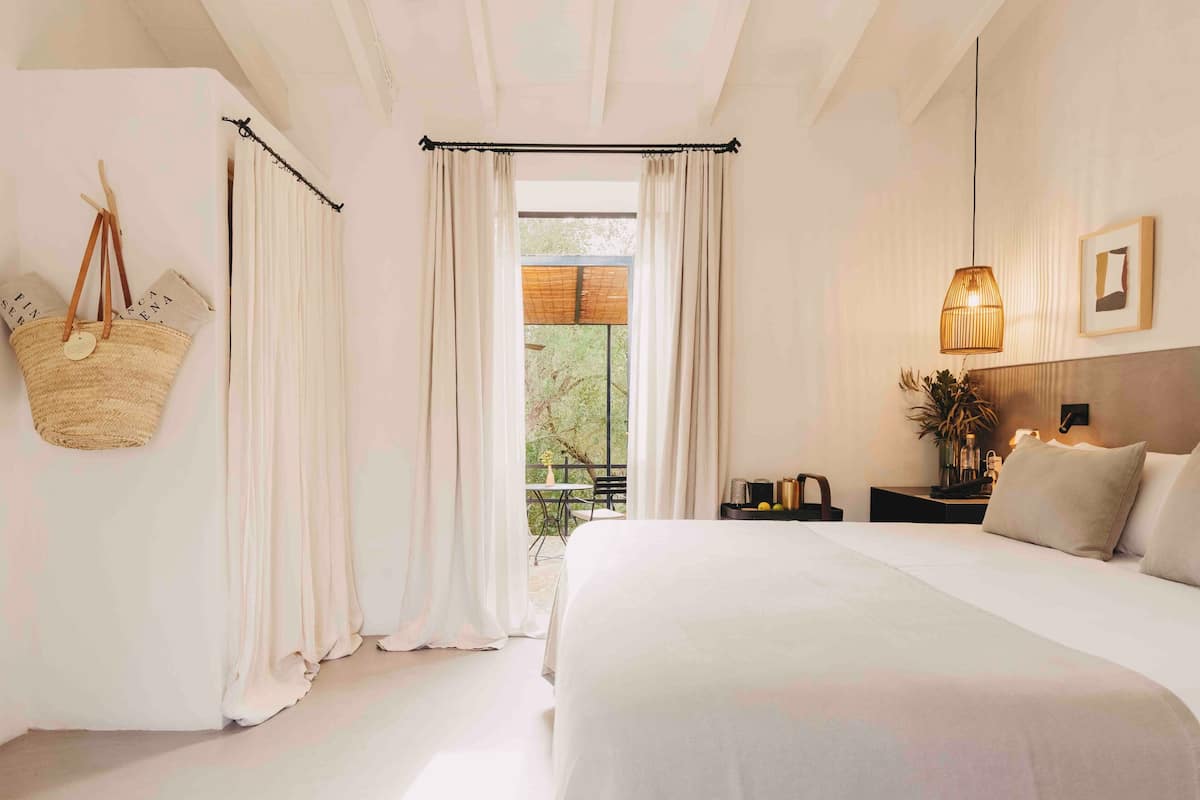 Ein Schlafzimmer mit einem großen Bett, einer hängenden Körbotage, einem Nachttisch mit Lampe und Blick ins Freie durch ein Fenster mit Vorhängen.
