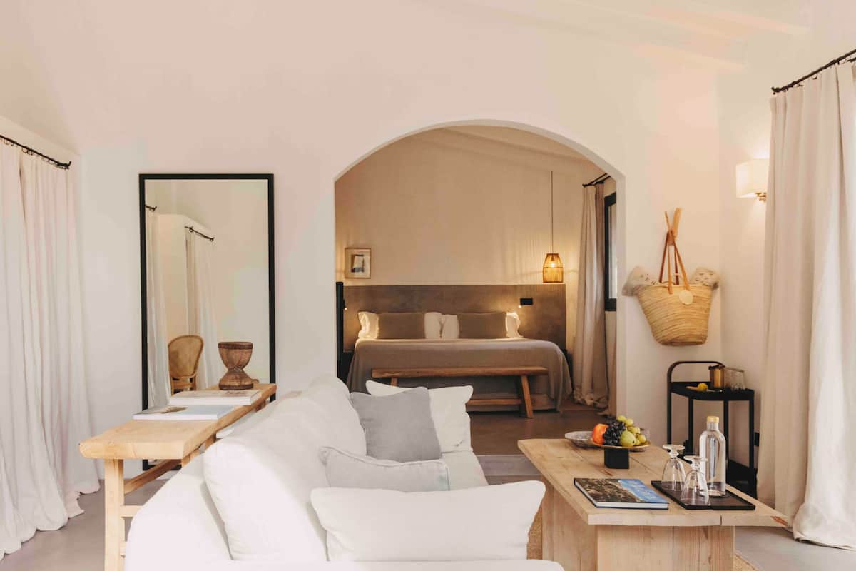 Ein modernes, minimalistisches Hotelzimmer mit einem großen Bett, einer Sitzecke mit einer Couch und einem kleinen Tisch sowie einem Esstisch mit Obstschale und Flasche.