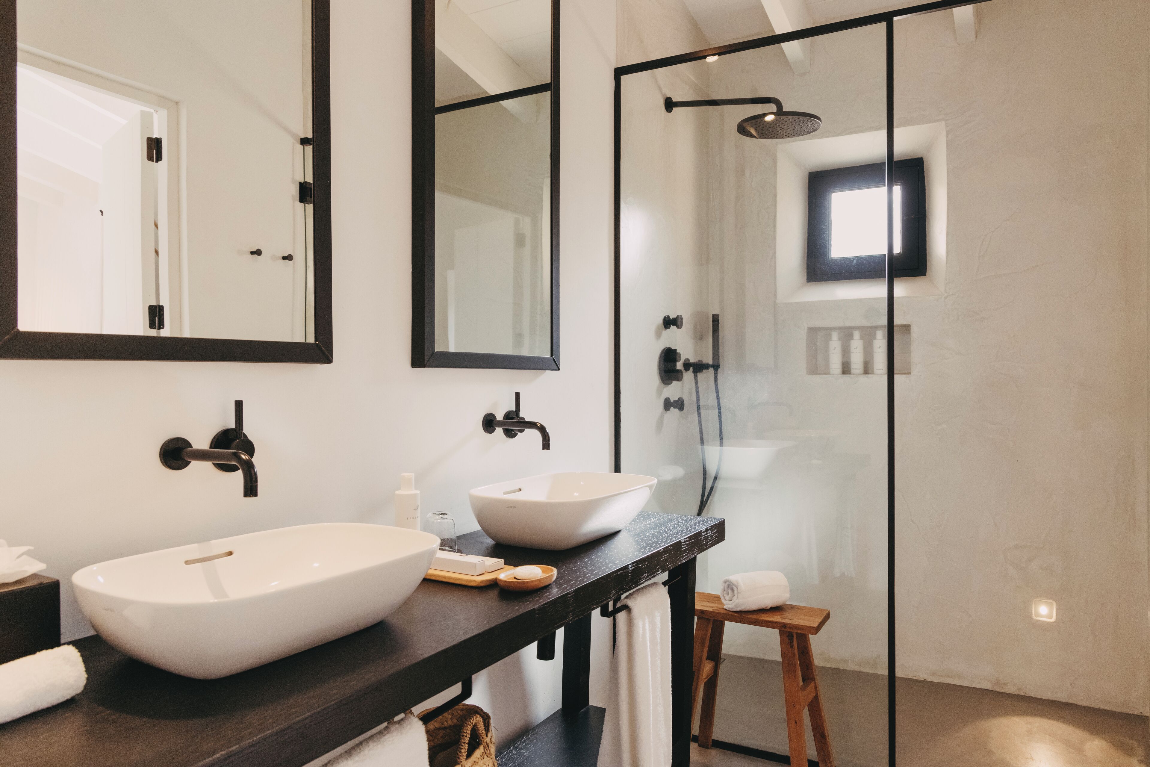 Un baño moderno con un lavamanos tipo recipiente grande, una encimera oscura, un taburete de madera y una zona de ducha con cerramiento de vidrio.