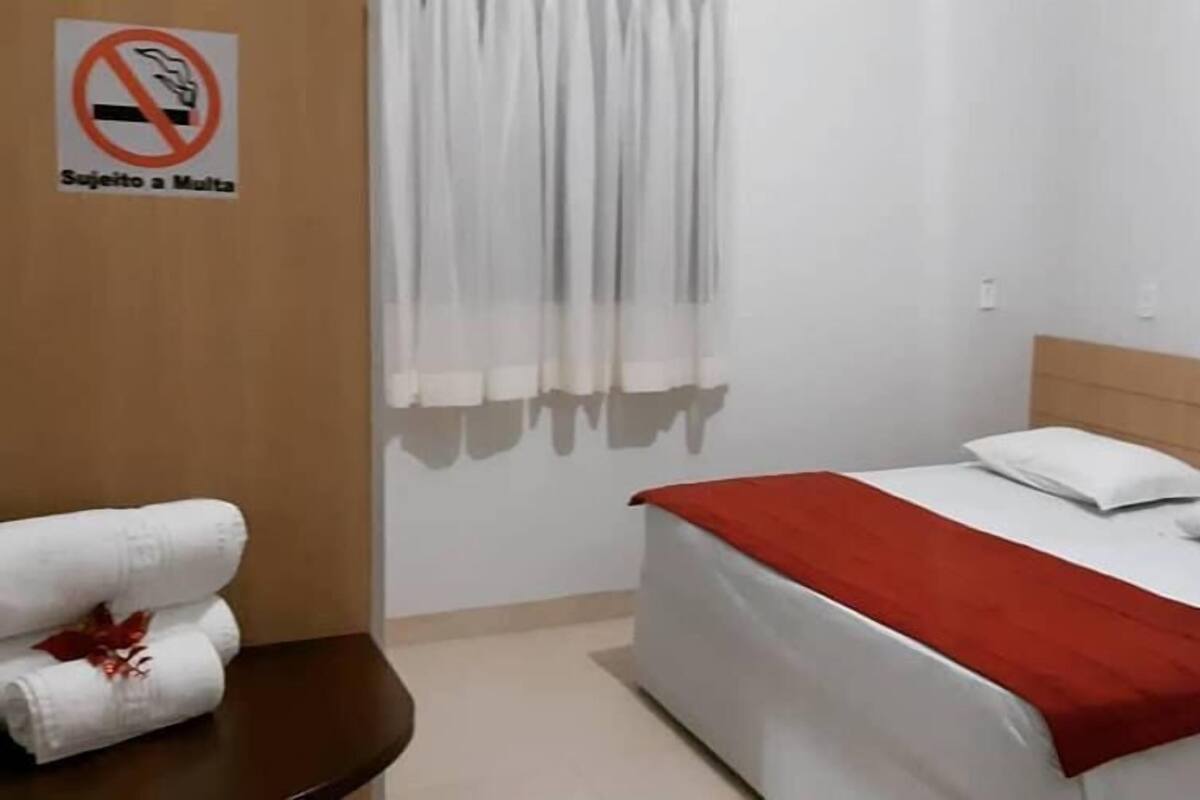 A hotel room with a bed, a red blanket, white pillows, a towel on a table, a no-smoking sign, and a painting on the wall.