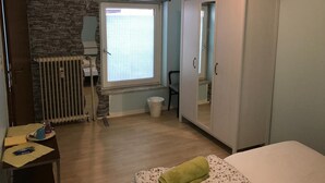 Una habitación con pared de piedra, una ventana con persianas, un armario, un radiador, una cama y una pequeña mesa con objetos encima.