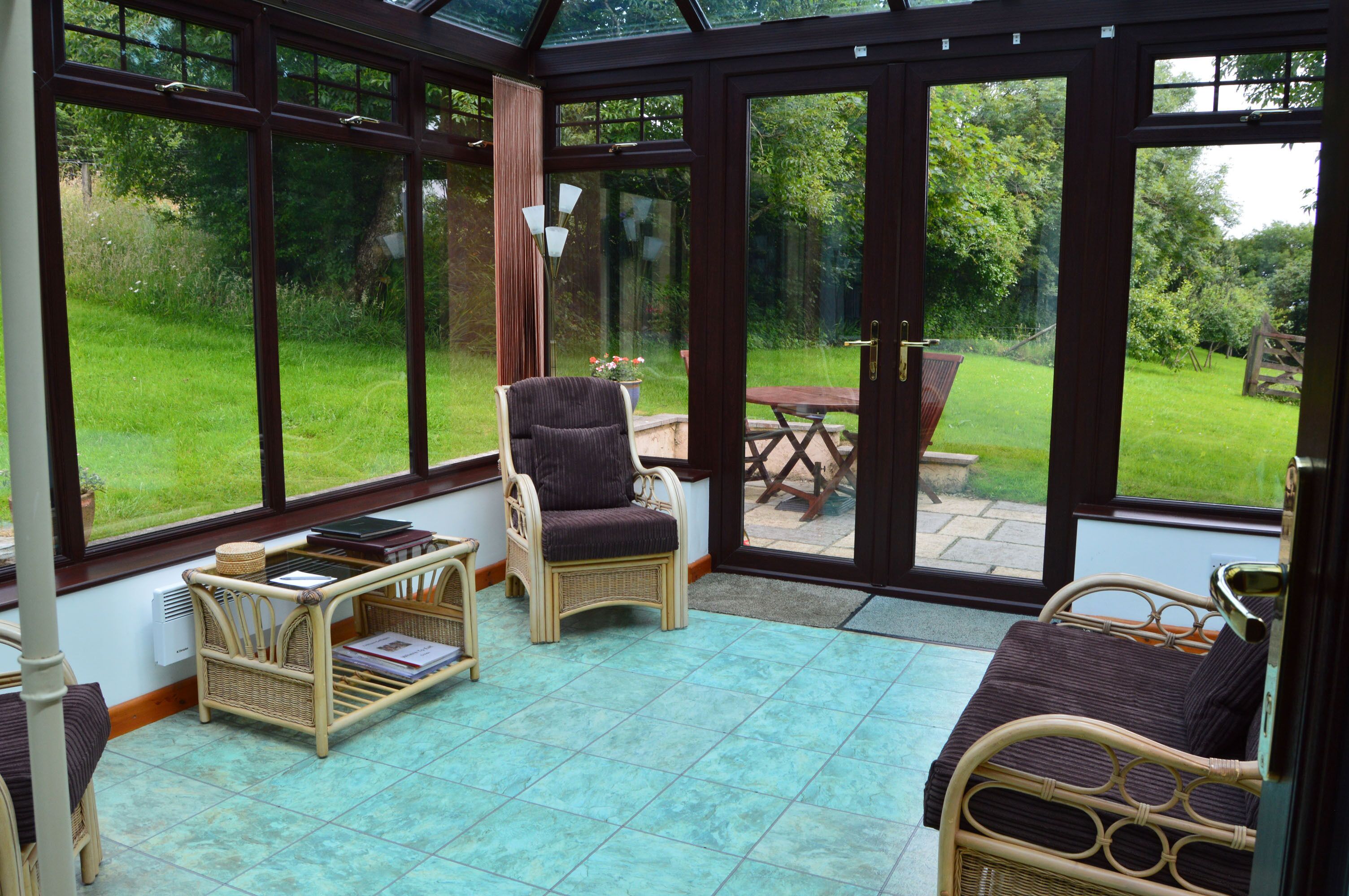 A conservatory with wicker furniture, a table, and chairs, overlooking a garden.