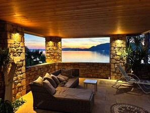 A covered outdoor seating area with stone walls, a wicker sofa, and a view of a lake at sunset.