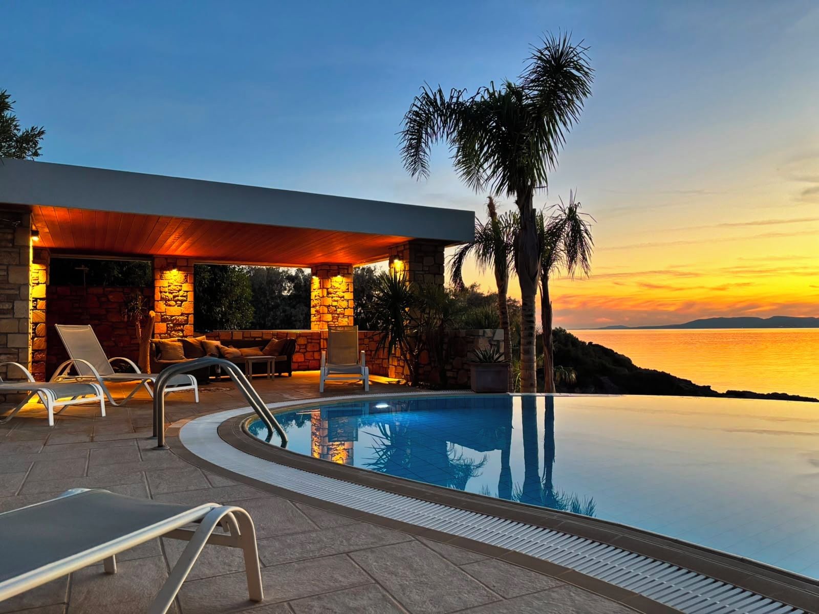 A poolside area with lounge chairs and a stone building, overlooking a body of water at sunset.