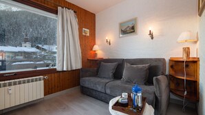 A cozy room with a wooden paneled wall, a grey sofa, a window with curtains, a radiator, and a small table with a tray. - Résidence le Chardonnet D Studio w/Shared Pool & panoramic views, Argentière (Chamonix), France (Argentière (Chamonix))