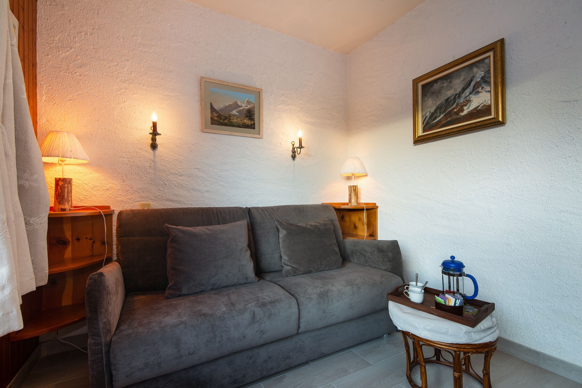 Un salon chaleureux avec un canapé gris, deux lampes fixées au mur, une petite table en bois sur laquelle se trouve une machine à café, et un tableau encadré accroché au mur.