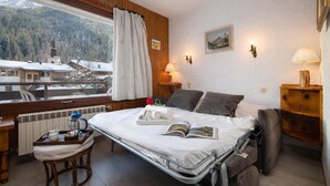 A hotel room with a bed, a small table with a kettle, a chair, a window with a view of a mountain and buildings, and a lamp on the wall. - Résidence le Chardonnet D Studio w/Shared Pool & panoramic views, Argentière (Chamonix), France (Argentière (Chamonix))