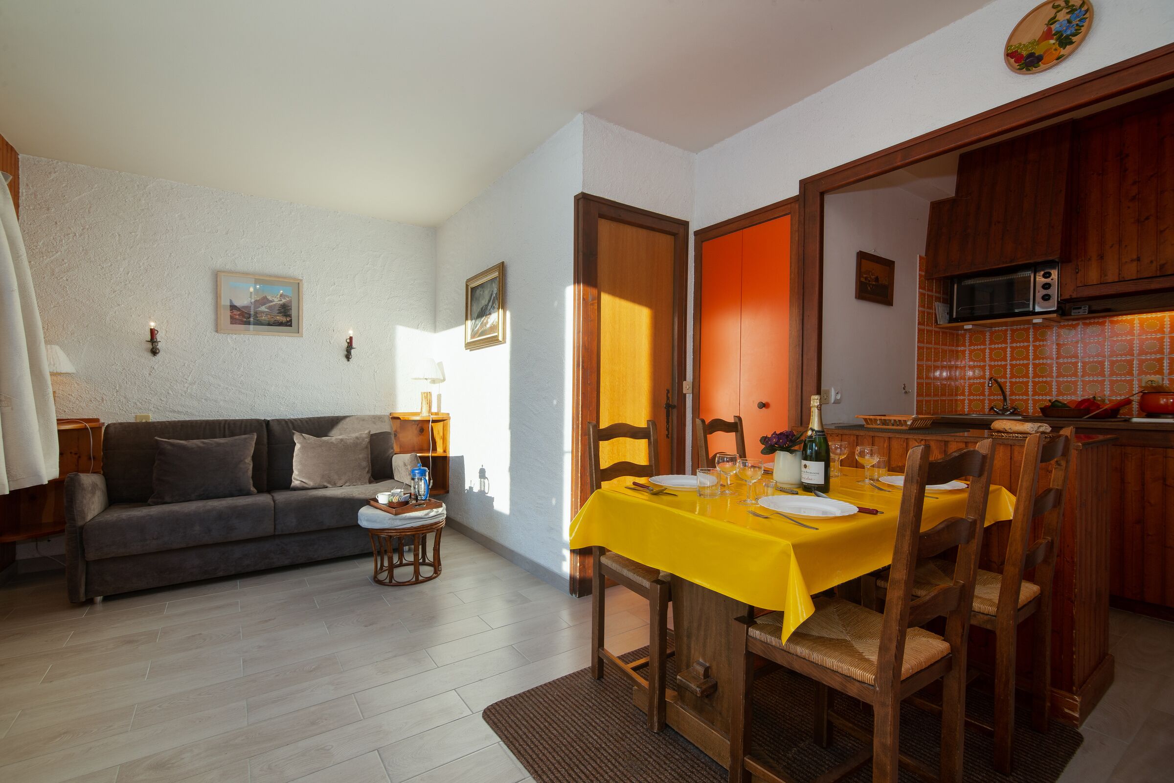 Un coin repas avec une nappe jaune, des chaises en bois et une cuisine visible au fond.