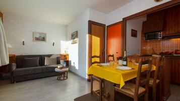Un coin repas avec une nappe jaune, des chaises en bois et une cuisine visible au fond.