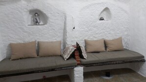 A cozy, white-walled room with a bench and cushions. There are two small shelves with decorative items.