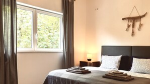 A bedroom with a large bed, a bedside table with a lamp, a window with curtains, and a wall-mounted decoration.