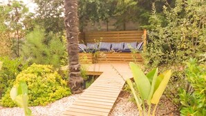 A wooden pathway leading to a bench with striped cushions, surrounded by lush greenery and plants.