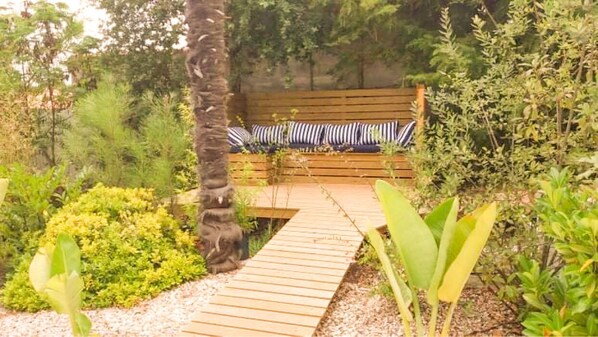 A wooden pathway leading to a bench with striped cushions, surrounded by lush greenery and plants.