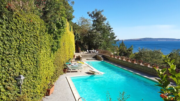Une piscine avec vue sur la mer, entourée de verdure et dotée d’un patio avec des espaces de détente en plein air.