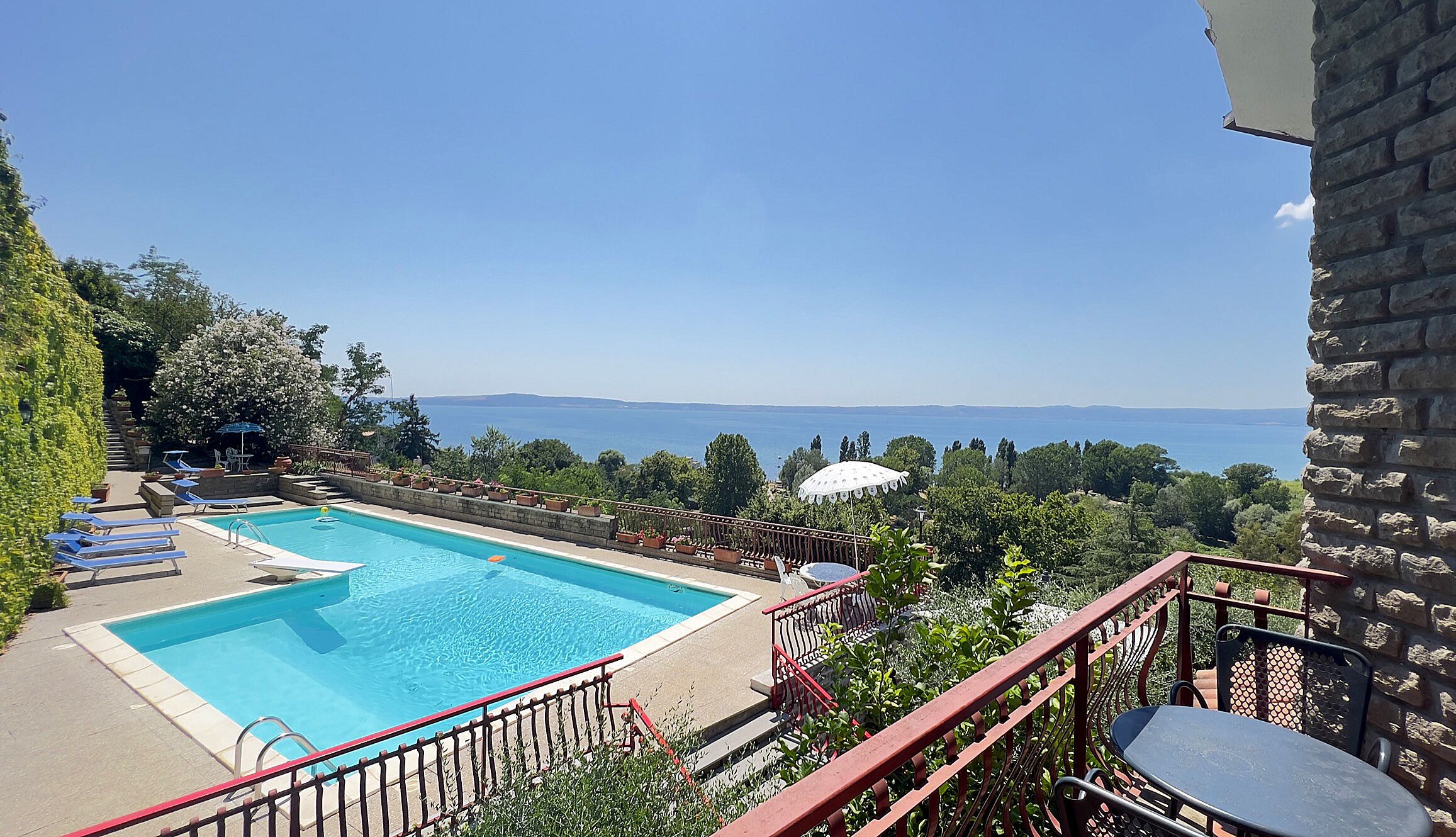 Une piscine avec vue sur la mer, entourée de verdure et bordée d’un mur de pierre.