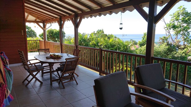 A covered outdoor patio with a wooden table and chairs, a view of the sea, and a railing.