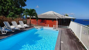 A swimming pool with white lounge chairs, a wooden deck, a red building with a white roof, and a view of the ocean.