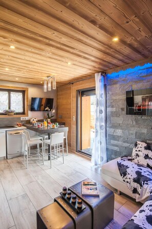 A modern kitchen with wooden ceiling, a dining area with white chairs, a sofa with a cow print cover, and a bar area with bottles. - NEW APARTMENT WITH PRIVATE JACUZZI CHEMINEE CLASS 5 STARS (Les Avanchers-Valmorel)