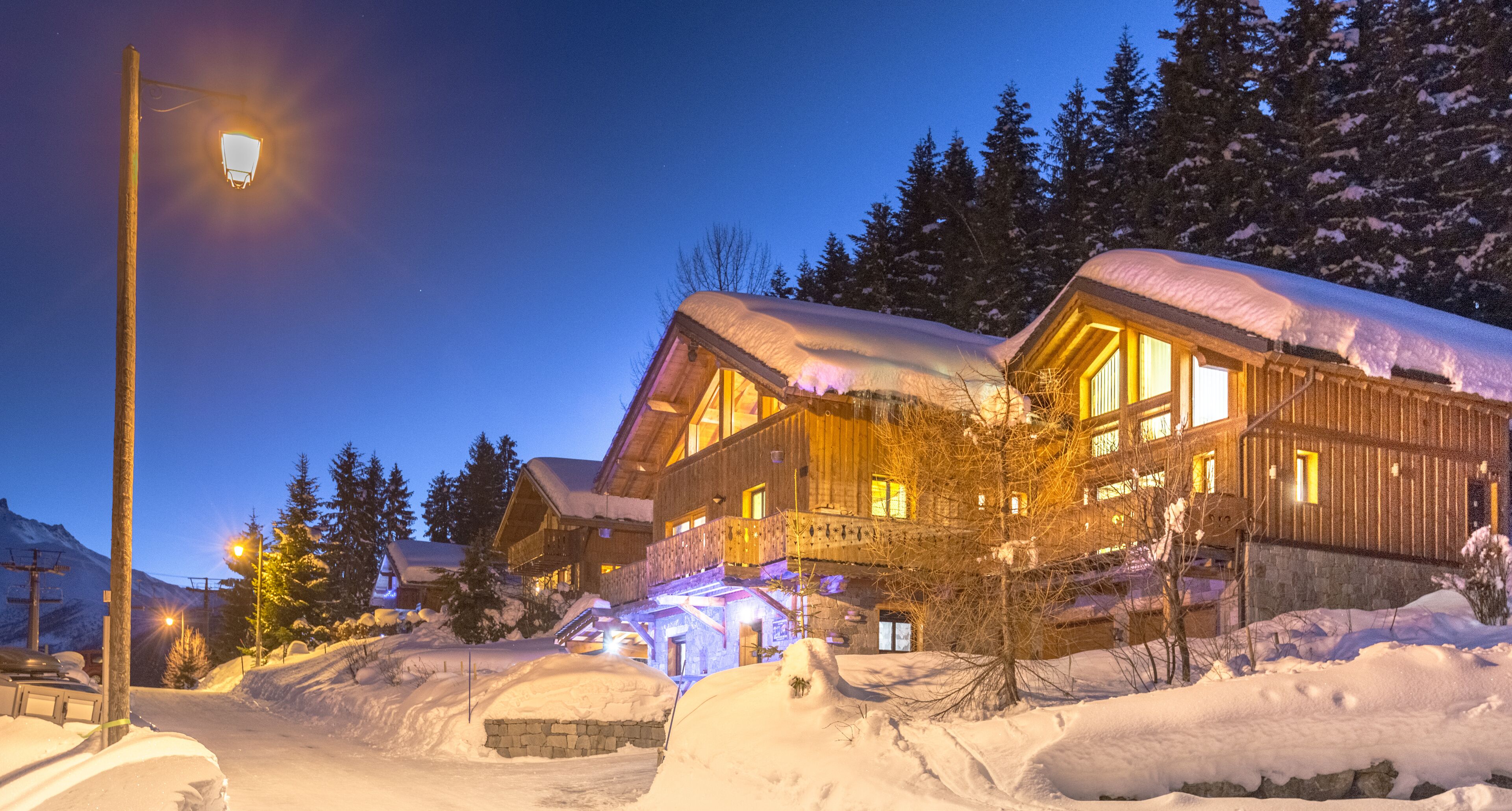 Un grand chalet en bois, avec un toit enneigé, des fenêtres illuminées et des fondations en pierre.