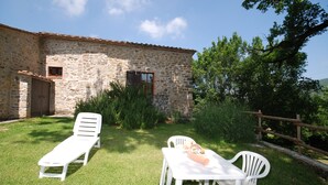 A stone building with a tiled roof, a small window, and a door. There is a white lounge chair and a table with a teddy bear on it in the foreground.