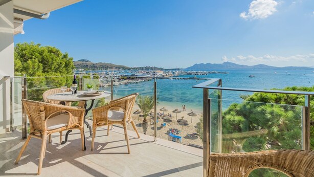 A balcony with wicker furniture, a table with a bottle and glasses, and a view of a beach and marina. - Apartment Colorado 3B By SunVillas Mallorca (Port de Pollensa)