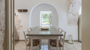 A dining area with a long wooden table, white chairs, and a view through an arched window to a garden.