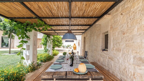 An outdoor dining area with a rustic wooden table, a thatched roof, and a stone wall.
