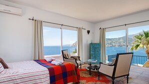 A bedroom with a bed, a chair, a small table, and a view of the sea.
