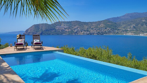 A swimming pool with two lounge chairs, a view of the sea, and a mountainous landscape in the background.