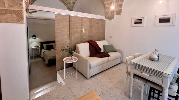 Un salon moderne avec un canapé blanc, une petite table ronde et une table à manger avec des chaises. On y trouve deux lampes suspendues et une plante posée sur une table d’appoint.
