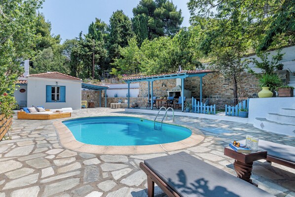 A pool area with stone flooring, a covered seating area, and a small building with blue shutters.
