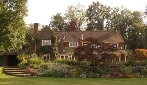 A large, two-story house with a brick exterior and multiple windows, surrounded by a well-maintained garden with various plants and flowers. - Private, spacious, elegant flat with lovely views, in a beautiful area (Westcott)