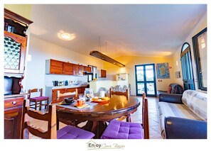 Amplia sala de estar con mesa de comedor, sillas y sofá. Se observa una cocina con armarios de madera y un ventanal grande.