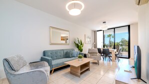 A modern living room with a grey sofa, a wooden coffee table, and a TV. There is a dining area with a table and chairs, and a large window offering a view of palm trees. - Oroklini - Perfectly overlooking the pool (Oroklini)