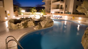 A swimming pool with a rocky edge, lounge chairs, and a poolside area with a view of a building in the background. - Oroklini - Perfectly overlooking the pool (Oroklini)