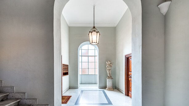 En hall med en marmortrappa, en stor, välvd dörröppning, en hängande lampa och en staty.