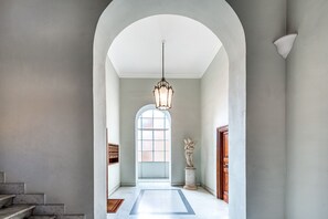 Un couloir avec un escalier en marbre, une grande porte en arc, un luminaire suspendu et une statue.