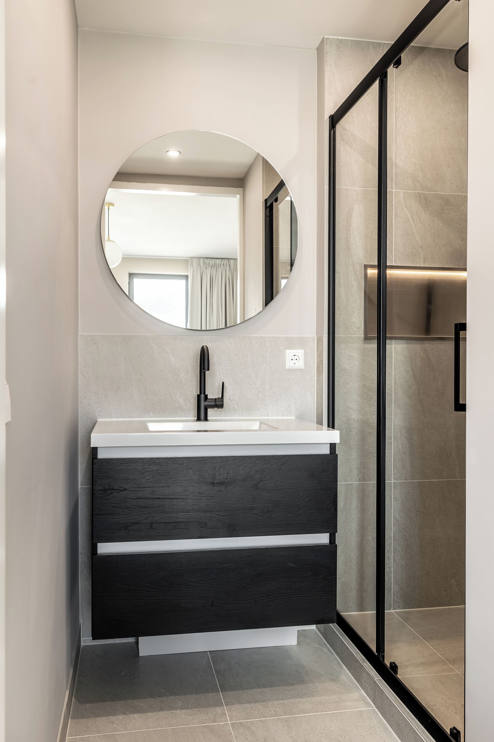 Een moderne badkamer met een ronde spiegel, een witte wastafel, donkerhouten kasten en een douchecabine met glazen wanden.