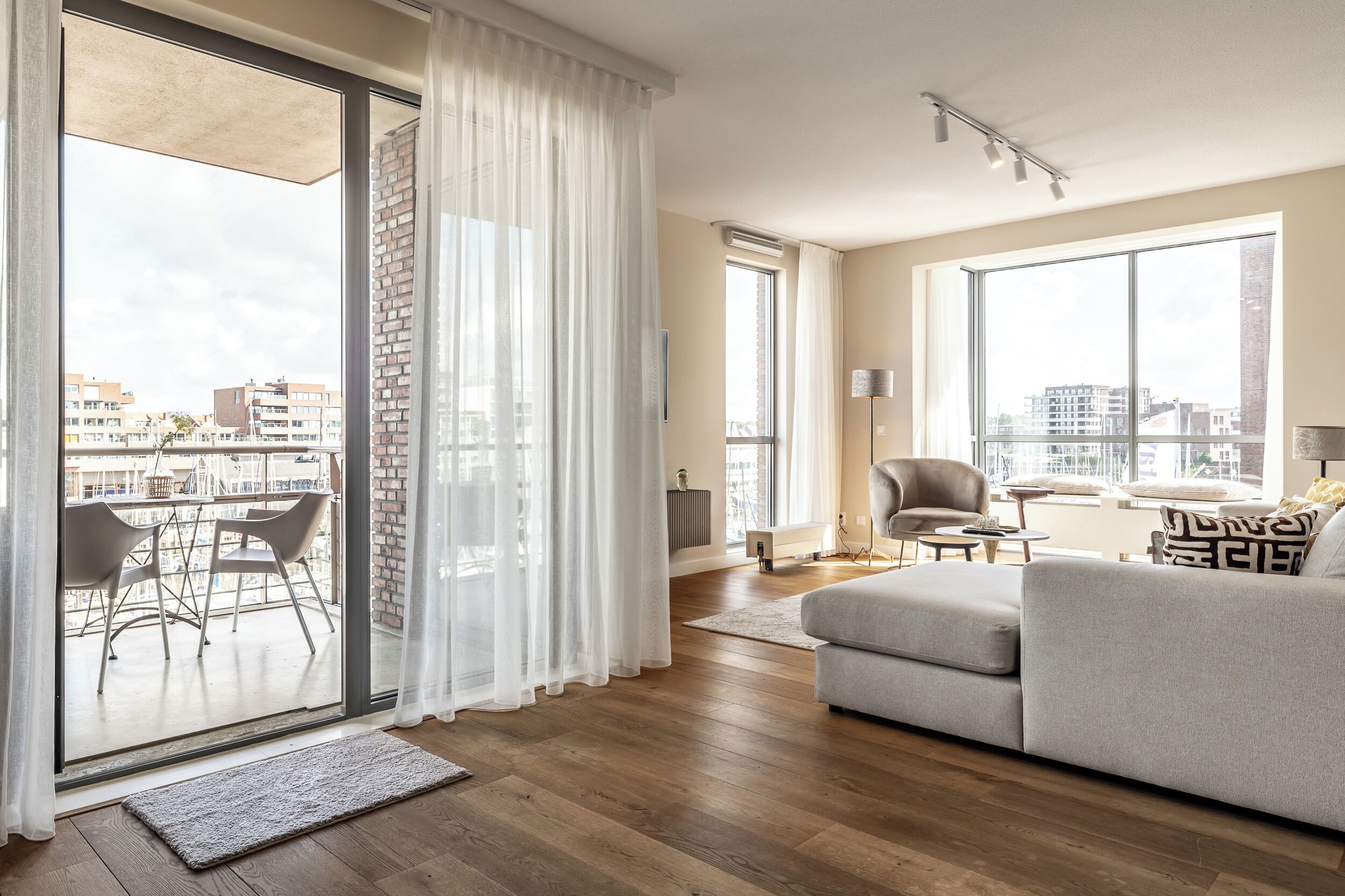 Una sala de estar moderna con un sofá gris, una mesa de comedor con sillas y un amplio ventanal corredero que da a un balcón exterior.