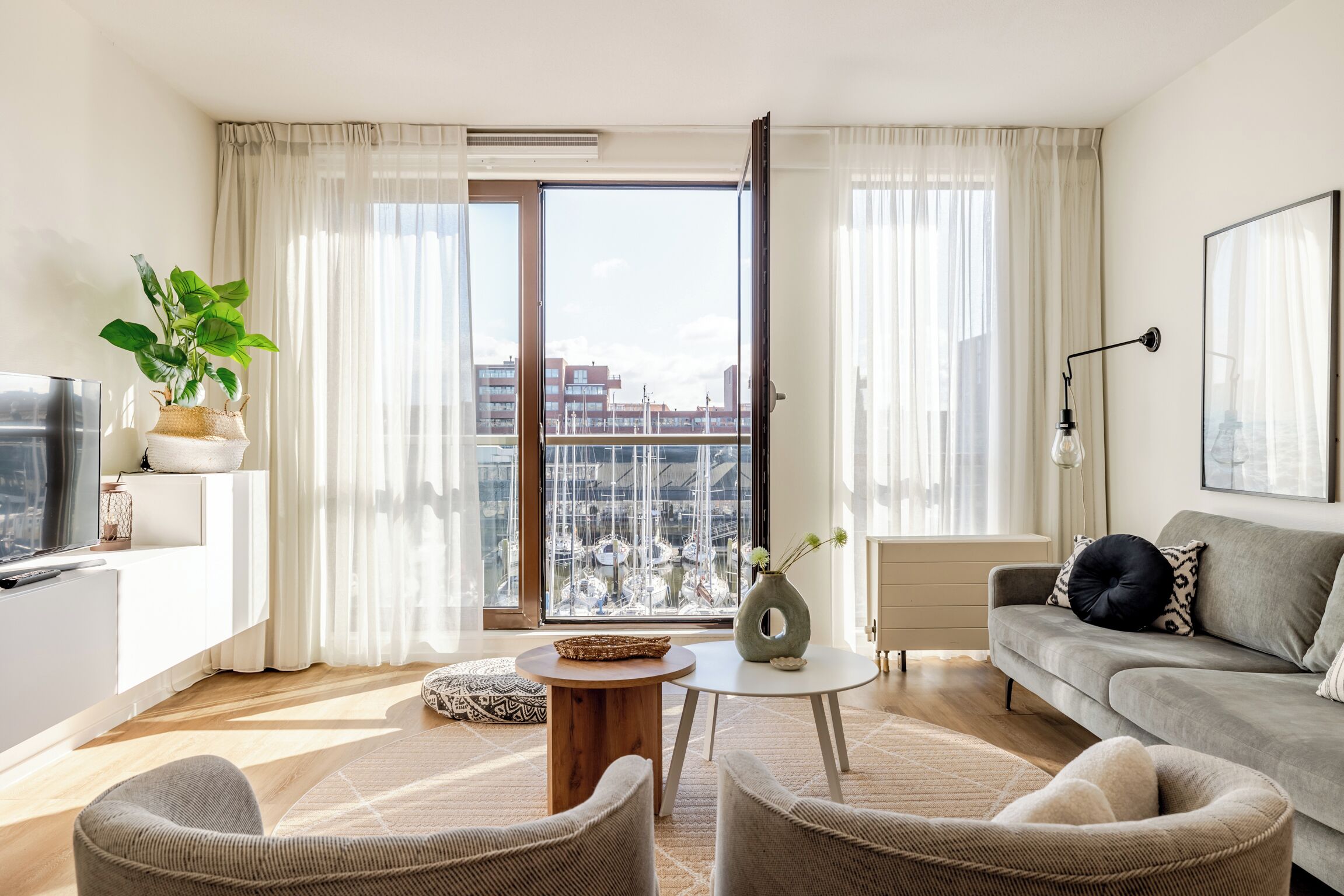 Un salon moderne comprenant un canapé, des fauteuils, une table basse, un téléviseur et une grande fenêtre donnant sur des immeubles et une marina.