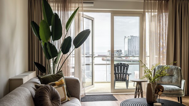 A modern living room with a sofa, a chair, a potted plant, and a view of the city through a glass door. - BizStay Harbour Scheveningen Apartments (The Hague)